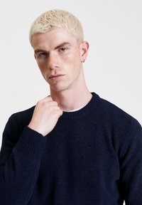 Granatowy sweter dzianinowy z okrągłym dekoltem, prążkowanymi mankietami i teksturowanym materiałem, noszony przez osobę, która trzyma się za kołnierz z neutralnym wyrazem twarzy.