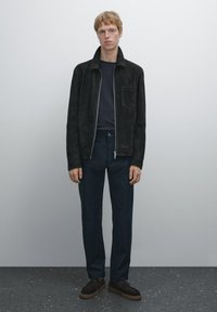 Černá semišová bunda se zipem a kapsou, v kombinaci s námořnickými kalhotami a tmavými šněrovacími botami, na neutrálním pozadí.