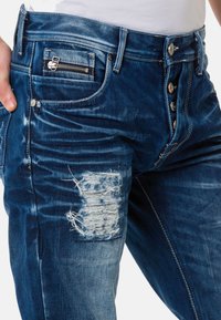 Dunkelblaue Denim-Jeans mit einem beschädigten Patch und Zipper-Tasche, ausgestattet mit einem Fünf-Knopf-Verschluss und dezentem Verblassen entlang der Beine.