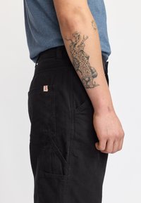 Pantalones cargo negros con una tela texturizada, que cuentan con múltiples bolsillos y una etiqueta roja en la parte trasera. Combinados con una camisa azul.