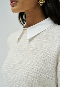 Pull en tissu crème texturé avec un col blanc. Présente des motifs subtils et une vue rapprochée du col et de l'épaule.