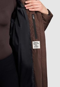 Ruskea tikattu ulkoverhoilu, jossa sileä musta vuori, sivussa vetoketjutasku ja etiketti, jossa lukee "Lämmin halaus kangasta."