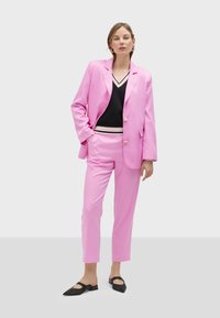 Pink blazer and matching trousers in smooth fabric, paired with a black top featuring a striped v-neck. Black flats complete the outfit.