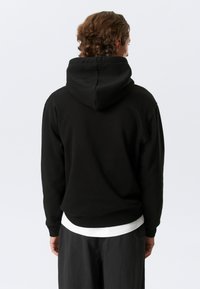 Must kapuutsiga dressipluus lõdva istuvusega, soonikkoes varrukaotsad ja ääred. Kannetud valge särgi peal, näidates lihtsat ja pehmet tekstuuri.