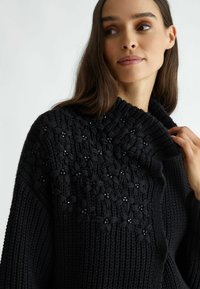 Černý pletený svetr s vysokým límcem, zdobený texturovanými květinovými vzory doplněnými korálky. Měkká, silná tkanina a uvolněný střih.
