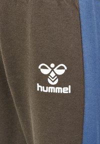Bruna träningsbyxor med en blå sidopanel och en vit "hummel"-logotyp som har ett bi-emblem, tillverkade av mjukt, elastiskt tyg.