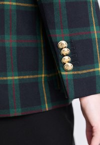 Manche de blazer à carreaux en bleu foncé, vert et jaune. Boutons dorés avec détails ornementaux. Tissu lisse avec une coupe structurée.