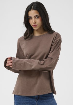 Femme aux longs cheveux foncés portant une chemise marron ample à manches longues et un jean bleu, debout, les mains jointes devant sur un fond blanc.