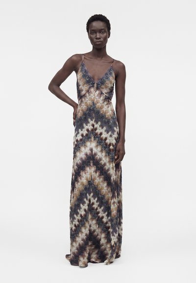 Ψηλή γυναίκα με κοντά σγουρά μαλλιά, που φοράει αμάνικο, μακρύ φόρεμα με V-λαιμόκοψη και ζιγκ ζαγκ μοτίβο σε γήινους τόνους.