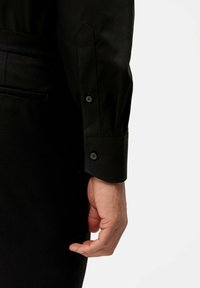 Schwarzes Langarmhemd mit geknöpften Manschetten und einem glatten, leicht strukturierten Stoff. Die Manschette hat eine spitze Form und zwei Knöpfe.