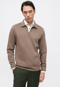 Sweater castanho texturizado com fecho, com colarinho, usado sobre uma camisa branca com detalhes em botão, combinado com calças verde-escuro.
