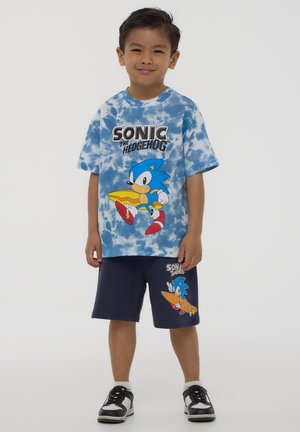 T-shirt blu con design tie-dye caratterizzato da una grafica di un riccio dei cartoni blu e la scritta "Sonic the Hedgehog", abbinato a pantaloni corti blu navy che mostrano un grafico simile.