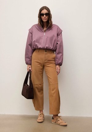 Femme portant une veste mauve à fermeture éclair, un pantalon large beige, des baskets beige, des lunettes de soleil noires, tenant un sac à main brun foncé, debout à l'intérieur.