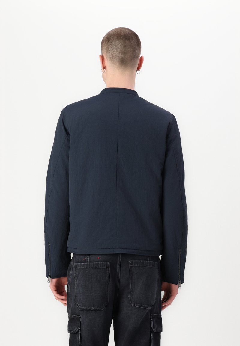 Veste bleu marine avec une texture lisse et un design minimaliste. Dotée d'un col montant et de poignets zippés. Portée par-dessus un pantalon cargo sombre.