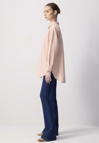 Ljusrosa långärmad skjorta med krage och knappmanschetter, bärs tillsammans med mörkblå utsvängda jeans. Slät tyg, avslappnad passform och längre bakstycke.