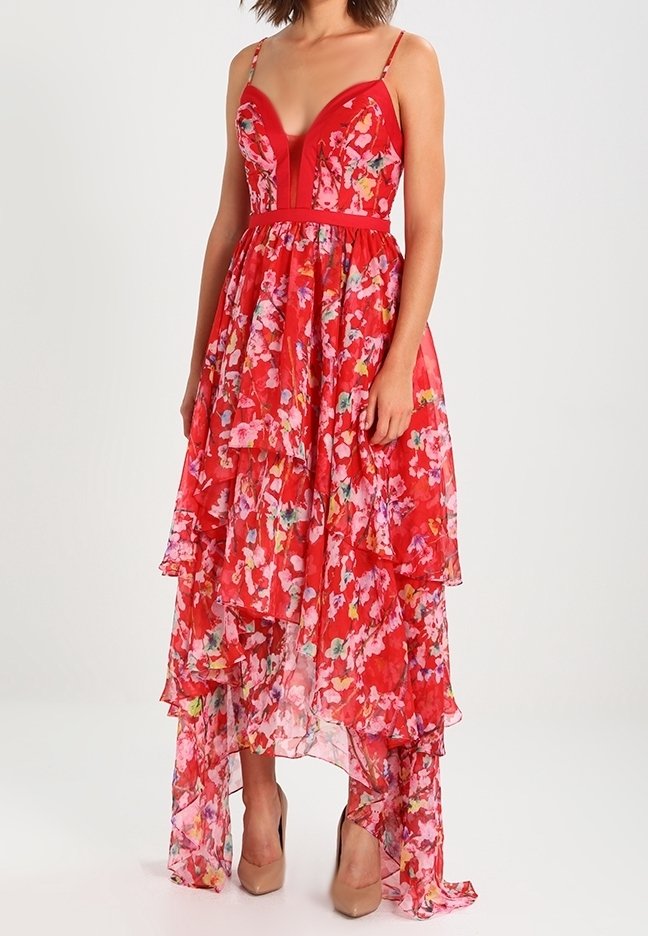 Femme portant une robe rouge fleurie à étages asymétrique avec fines bretelles et des talons hauts couleur nude, debout devant un fond uni.
