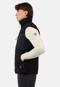 Gilet in fleece nero con alto colletto, pannello in tessuto liscio in contrasto, tasche laterali con zip e badge con logo sul lato sinistro del petto.