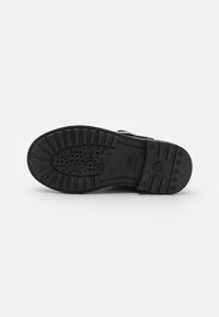 Suela de zapato de goma negra con patrón texturizado de agarre, perforaciones circulares y detalle del logo. Diseño plano con secciones elevadas para tracción.
