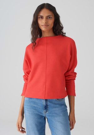 Maglione rosso a maniche lunghe con una vestibilità rilassata, caratterizzato da un collo a barca e una cucitura centrale, abbinato a jeans azzurri chiari. Texture liscia.