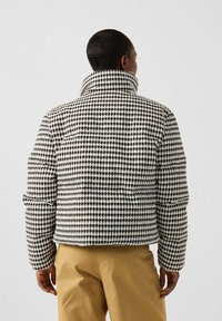 Persoon met kort haar, gekleed in een zwart-wit houndstooth patroon jas en beige broek, met de rug naar de camera tegen een effen achtergrond.