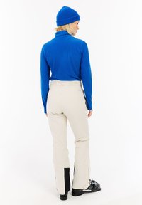 Blauer gerippter Rollkragenpullover, beigefarbene Skihose mit schwarzen Akzenten und schwarze Skischuhe. Von hinten betrachtet mit einem figurbetonten Design.