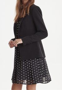 Czarna, dopasowana marynarka na jeden guzik, noszona na czarnej sukience w groszki. Sukienka ma plisowaną spódnicę i przezroczystą tkaninę.