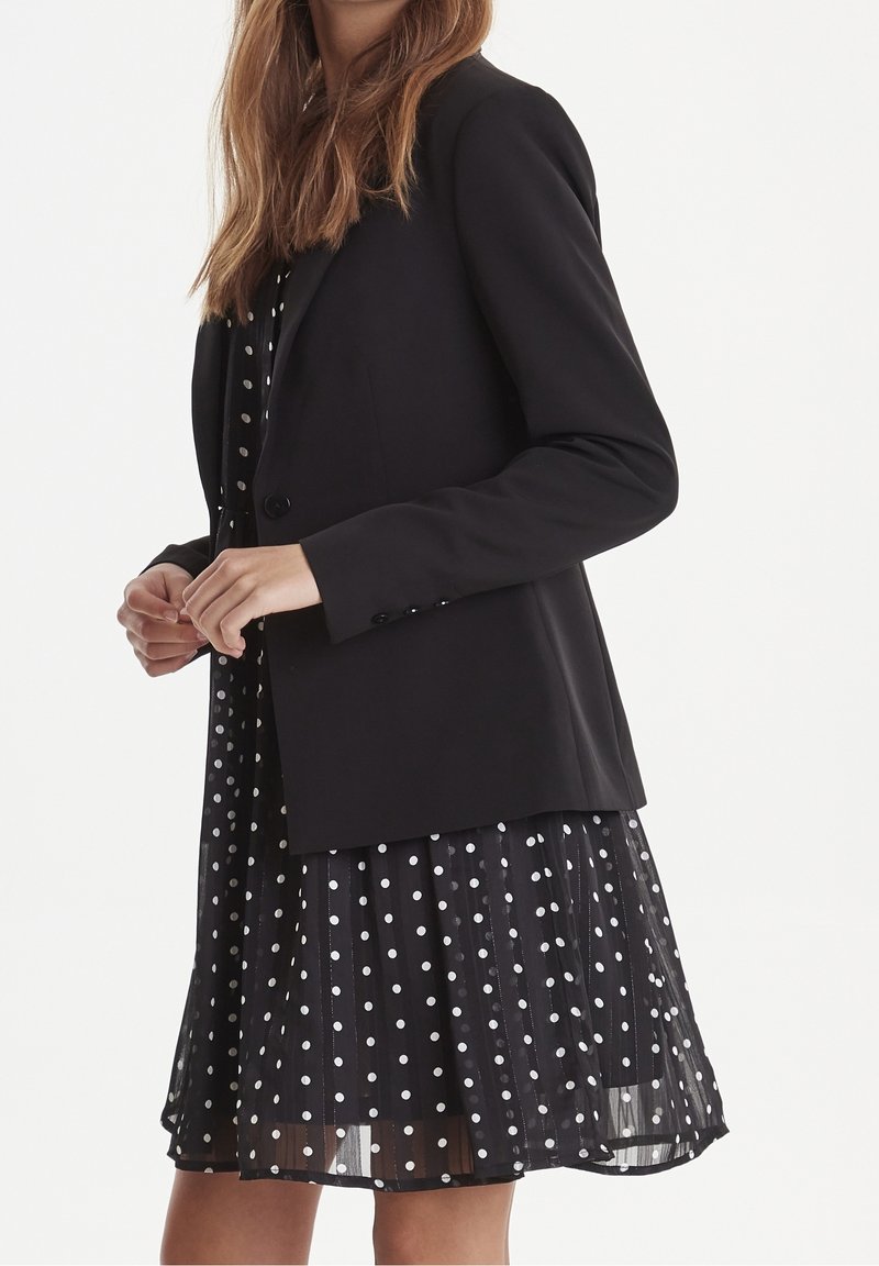 Czarna, dopasowana marynarka na jeden guzik, noszona na czarnej sukience w groszki. Sukienka ma plisowaną spódnicę i przezroczystą tkaninę.