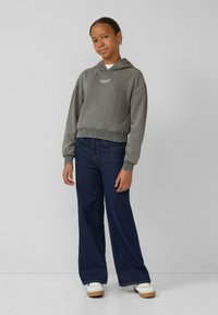 Adolescente vestido con una sudadera gris deslucida con texto, jeans anchos oscuros y zapatillas blancas, de pie contra un fondo liso gris claro.