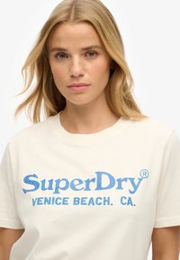 Kremowy bawełniany t-shirt z niebieskim tekstem z cyrkonii „SuperDry VENICE BEACH. CA.” Krótkie rękawy i klasyczny okrągły dekolt.