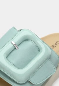 Σανδάλι πλάγιο σε μέντα πράσινο, με μεγάλο γυαλιστερό αγκράφα, λεία υφή δέρματος και επένδυση με μαξιλάρι. Τα μεταλλικά μέρη είναι ασημί χρώματος.