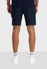 Pantalones cortos azul marino hechos de una tela elástica con acentos blancos a los lados. Llevados con zapatillas blancas y calcetas deportivas. Textura suave, diseño ajustado.