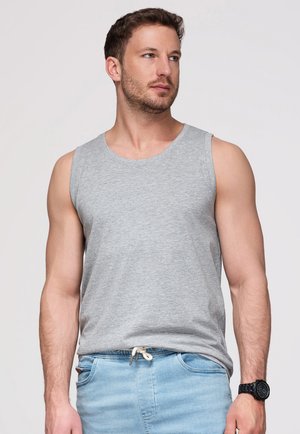 Homme portant un débardeur gris clair et un jean bleu clair avec une montre, regardant à droite sur un fond uni.