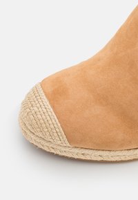 Tan mockasiner i mocka med rund tå och en vävd juteaccent vid tåhuvudet. Texturerad yta och detaljer av naturlig fiber.