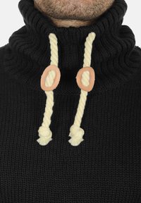 Jersey negro de punto con cuello alto, que presenta cordones trenzados de cuerda de color crema y detalles circulares en tono camel. Patrón texturizado en el cuello.