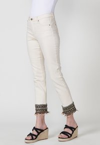 Jeans beige hasta los tobillos con un puño decorativo bordado que incluye cuentas, complementados por sandalias de cuña negras con tiras.