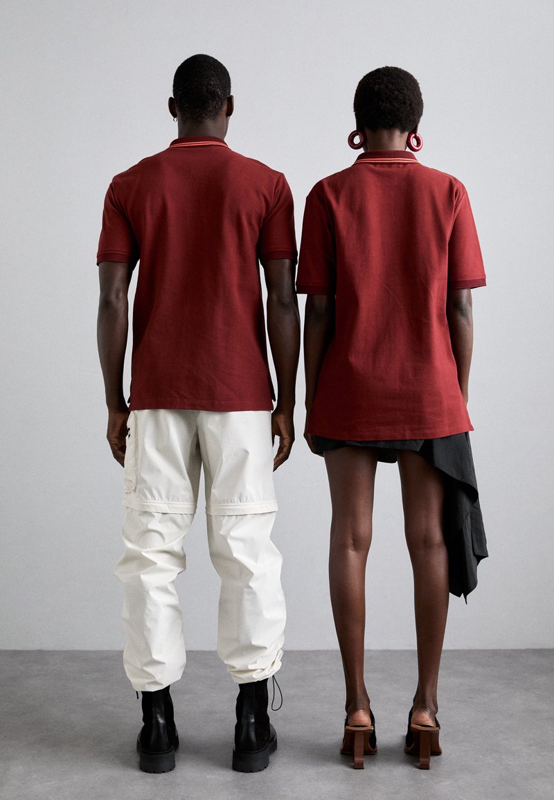 Twee personen staan rug aan rug. Beiden dragen vergelijkbare bordeauxrode polo shirts. De een draagt het met witte cargobroek, de ander met een zwart asymmetrische rok.