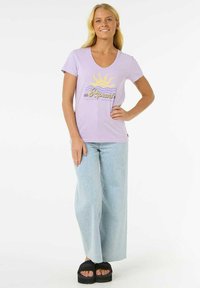 Lilla v-hals t-shirt med gul solgrafik og tekst, parret med lyseblå brede jeans og sorte platform-sandaler.