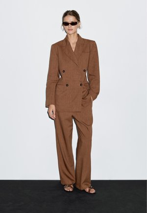 Bruine linnen dubbele-breasted blazer met twee voorzakken, gecombineerd met bijpassende wide-leg broek. Model draagt slanke zonnebril en sandalen.