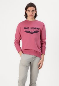 Różowy sweter z czarnym logo 'PME Legend American Classic', wykończony ściągaczami przy rękawach i u dołu. Model nosi jasnoszare spodnie i stoi z dłońmi w kieszeniach.