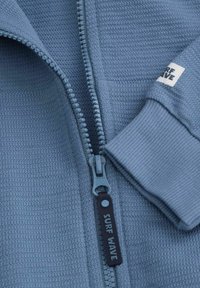 Jasnoniebieska kurtka zapinana na zamek, wykonana z teksturowanej tkaniny. Posiada gładki zamek błyskawiczny oraz ściągacze na rękawach z małą metką z napisem "SURF WAVE."