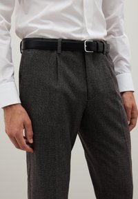 Pantaloni grigi con texture e un motivo sottile, abbinati a una cintura in pelle nera con fibbia argentata e una camicia bianca con colletto.