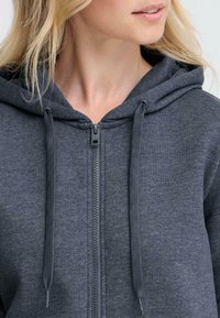 Grå zip-up hoodie med snoretræk i hætten, der har en glat, blød tekstur og synlige sømme langs lynlåsen og kanten.
