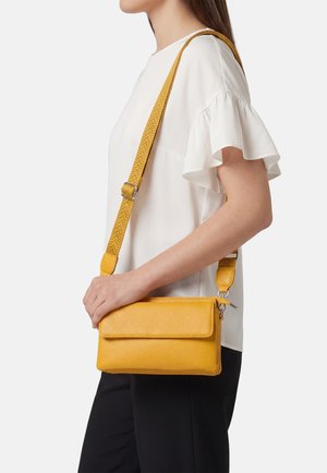 Donna con camicetta bianca e pantaloni neri tiene una borsa a tracolla in pelle gialla con tracolla regolabile sulla spalla.
