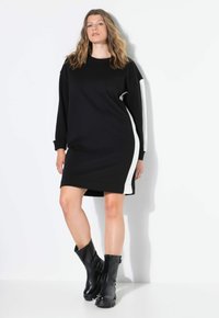 Vestido sudadera negro con franjas laterales blancas, mangas largas y puños acanalados. Combinado con botas de cuero negras hasta el tobillo sobre un fondo liso.