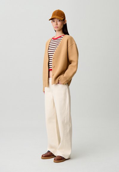 Cárdigan beige de punto con grandes botones, camisa a rayas rojas y blancas, pantalones crema sueltos y zapatos marrones con cordones. Gorra marrón completa el conjunto.