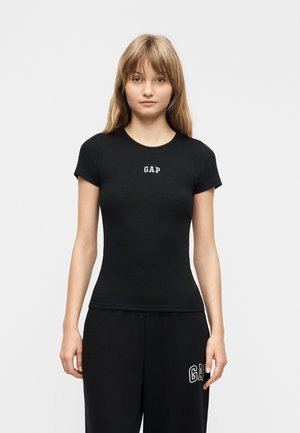 Jeune femme debout face à l'avant, portant un t-shirt noir ajusté de la marque GAP et un pantalon noir avec le logo GAP sur la cuisse gauche.