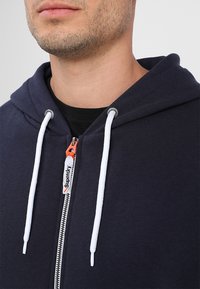 Námořnická mikina na zip s bílými stahovacími šňůrkami, s oranžovým lístkem na zipu a černou vnitřní podšívkou. Měkká látka s žebrovanými manžetami.