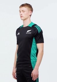 Camiseta atlética negra con paneles laterales verdes, el logo de los All Blacks y tres rayas verdes en los hombros. Diseño ajustado y de manga corta.