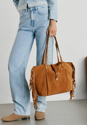 Isik, kes kannab helesiniseid teksaseid tikitud külje mustriga, hoiab suurt pruuni seemisnahast käekotti, millel on narmad ja pandla detail.