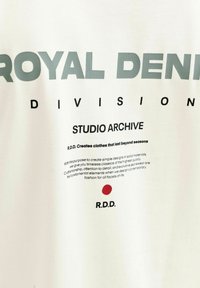 Weißer Stoff mit Text: ROYAL DENIM DIVISION, STUDIO ARCHIVE, Slogan auf Qualitätskleidung, roter Punkt über dem R.D.D.-Logo.